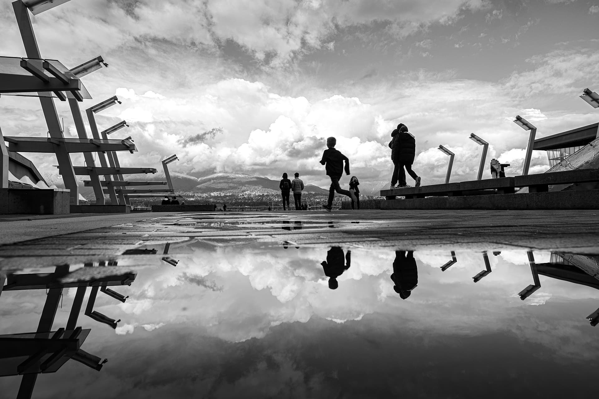 Silhouetted figures at the Vancouver Convention Centre plaza mirrored in a puddle under a dramatic cloud-filled sky, 16mm reflection street photography.