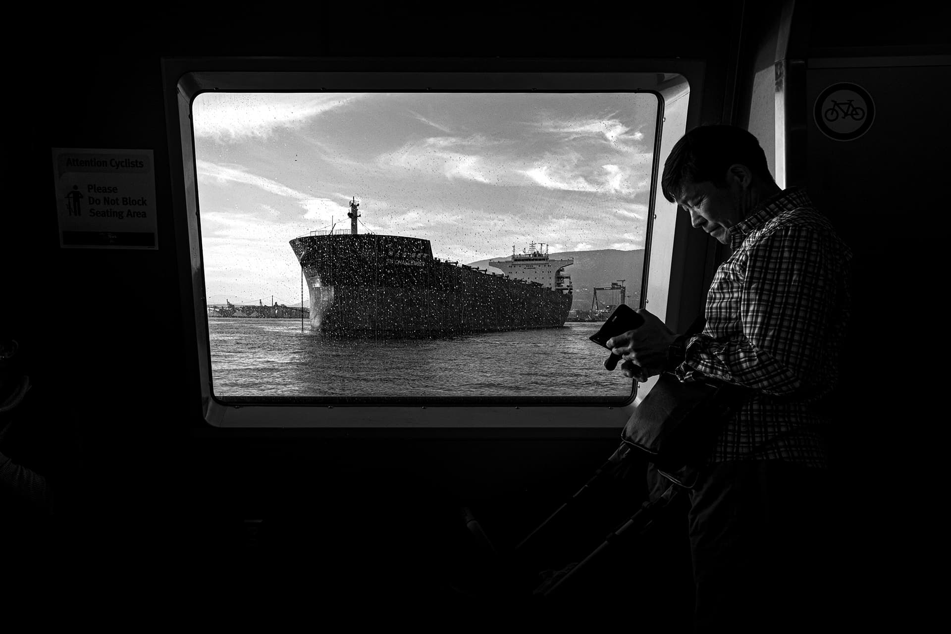 Passenger on a Vancouver SeaBus checking his phone as a cargo ship passes through the rain-spotted window behind him, candid black and white street photography.