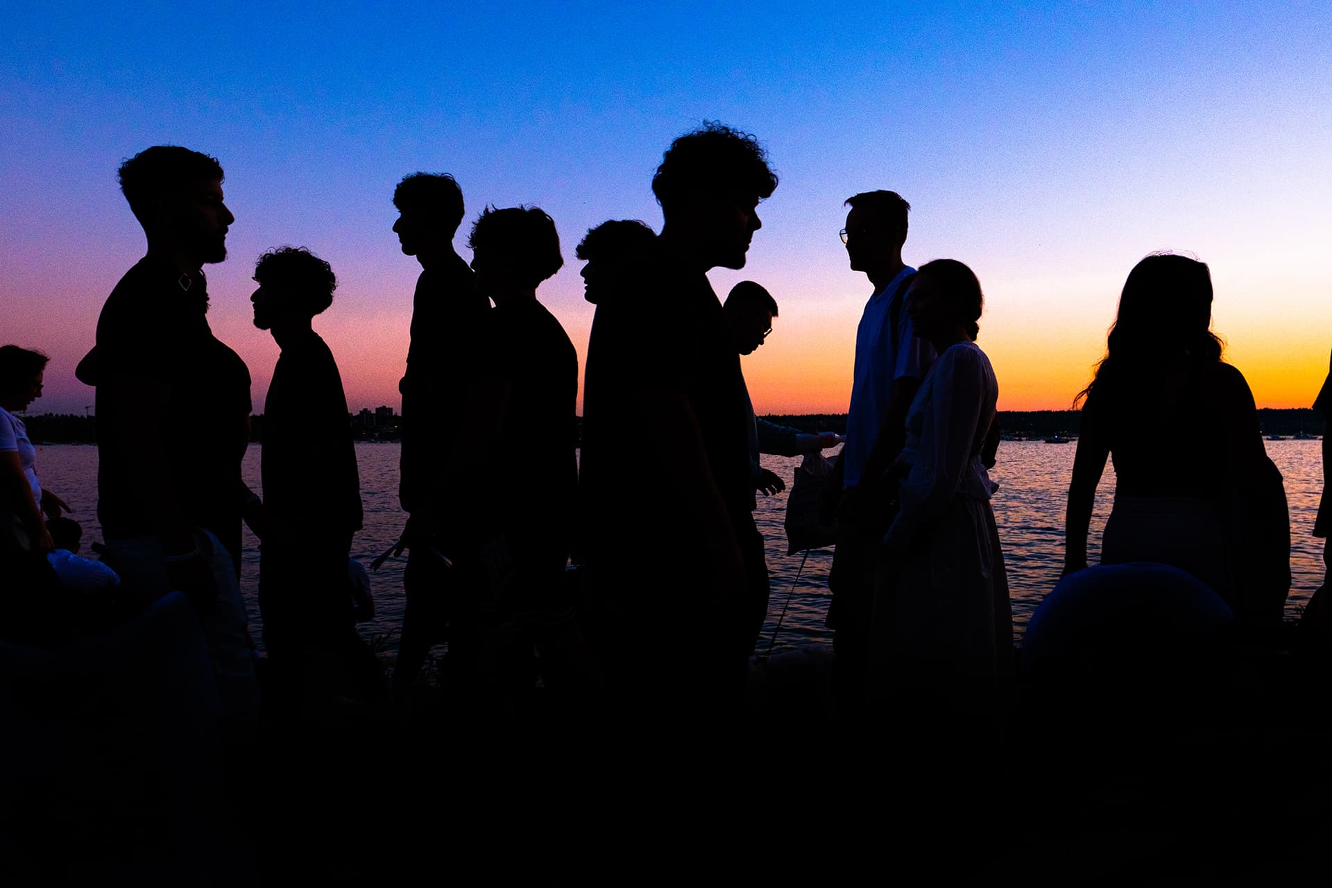 Group of silhouetted friends gathered at a Vancouver seawall under a blue-to-orange gradient sunset, colour street photography.